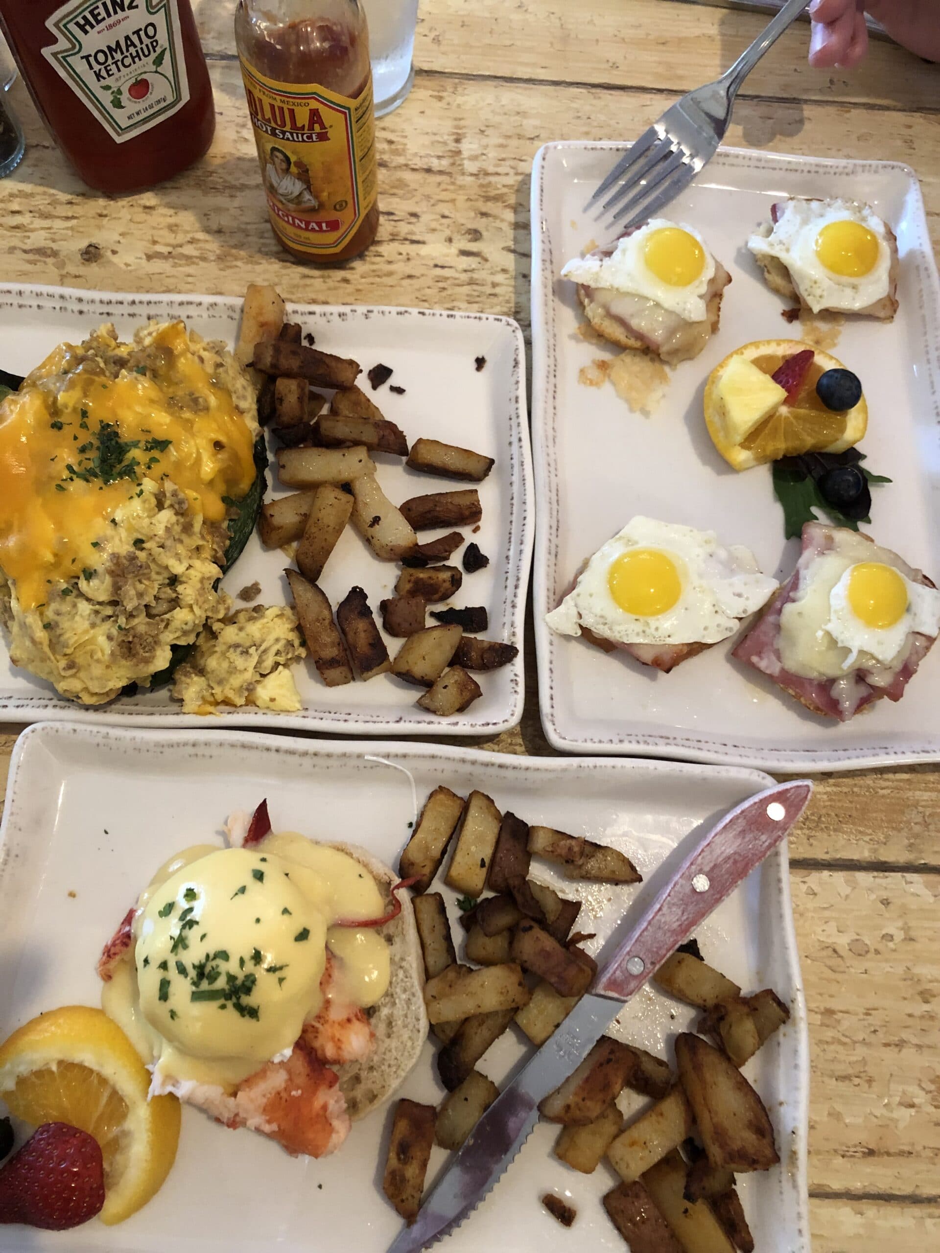 A plate of scrambled eggs with cheese, breakfast potatoes, and two dishes of eggs Benedict garnished with fruit.
