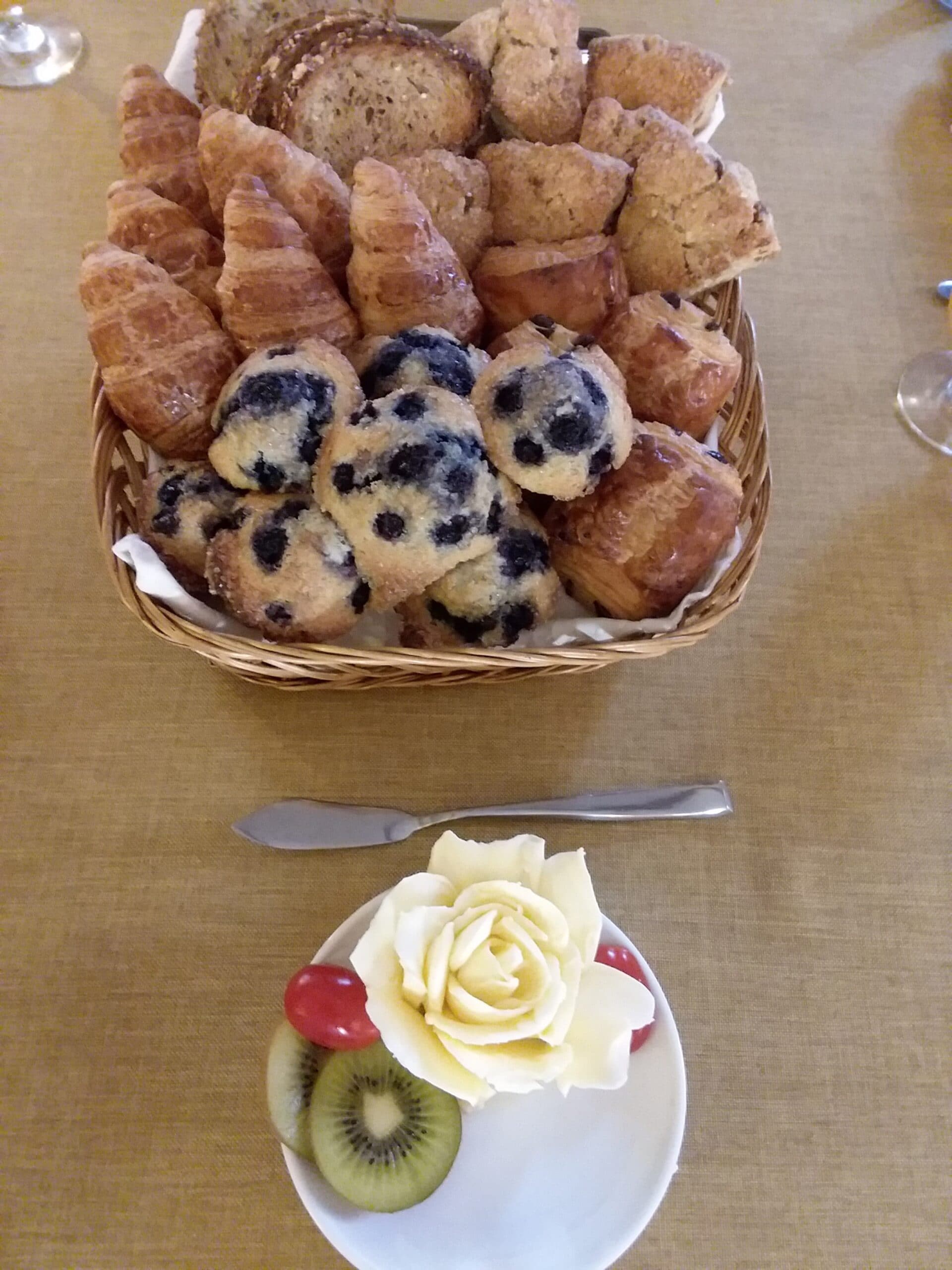 A basket of assorted pastries including croissants and blueberry muffins next to a plate featuring a decorative fruit arrangement.
