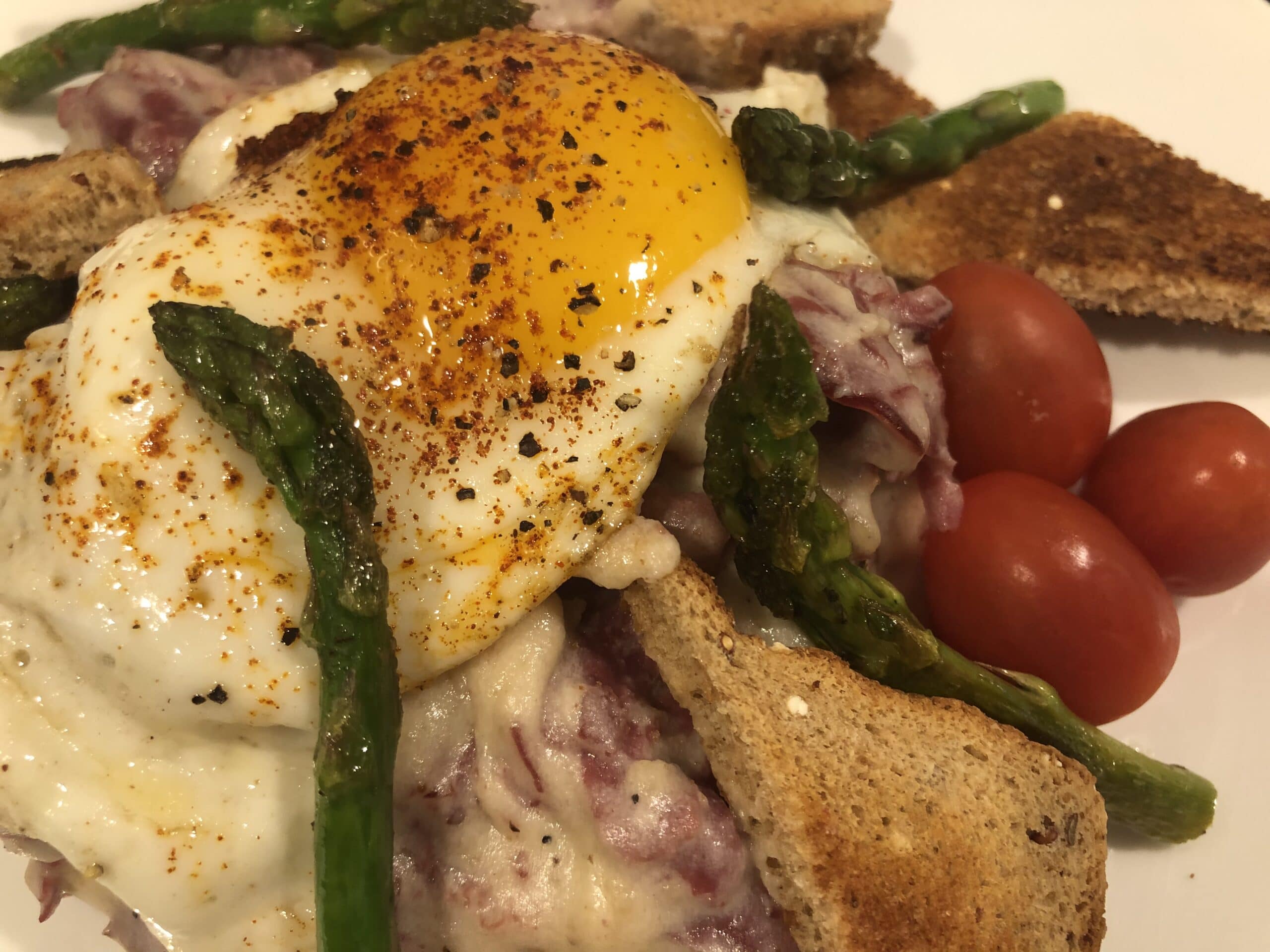 A close-up of a poached egg on creamy sauce with asparagus, toasted bread, and cherry tomatoes.