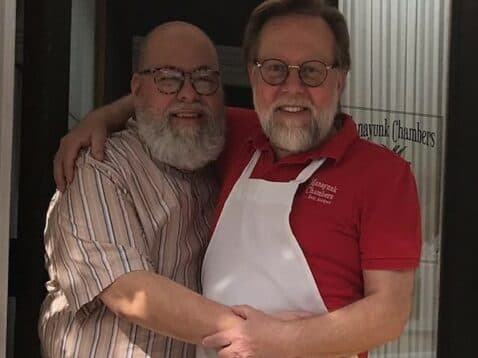 Two smiling men embrace warmly outside a door, one wearing a red shirt and an apron.