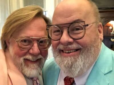 Two smiling men with glasses and beards pose closely together, both dressed in colorful attire.